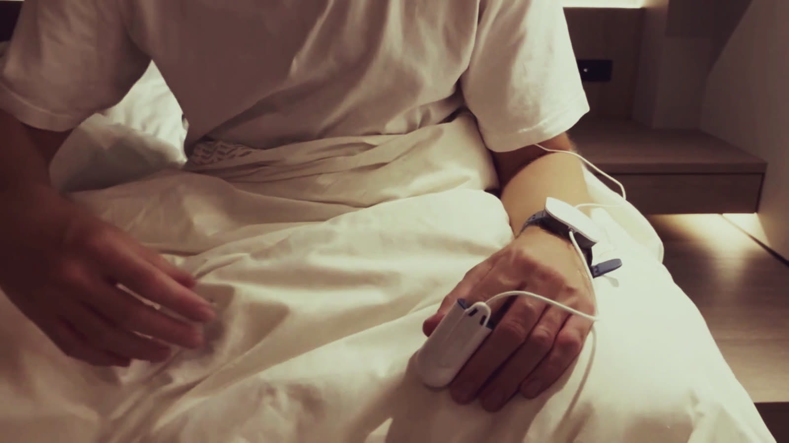 Iemand zit op de rand van het eigen bed en doet de vingersensor om; de polsunit zit al om. Warm lamplicht, gezicht buiten beeld.