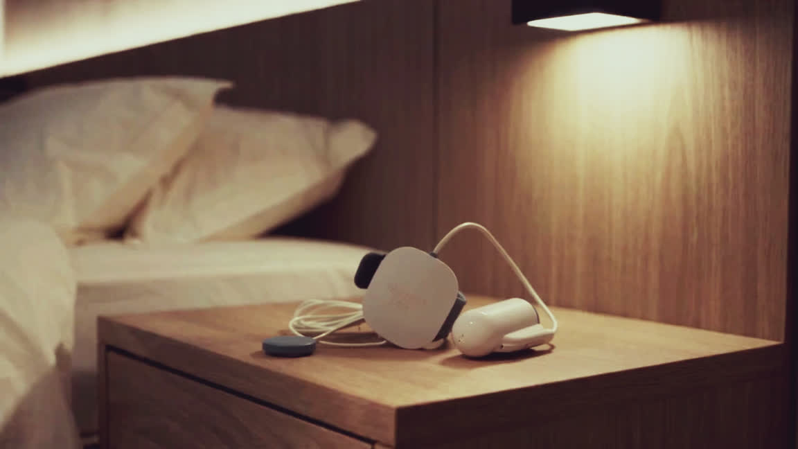 De drie delen van de thuis-slaaptest liggen klaar op een nachtkastje; daarna doet iemand op de rand van het bed de vingersensor om. Warm lamplicht, gezicht buiten beeld.